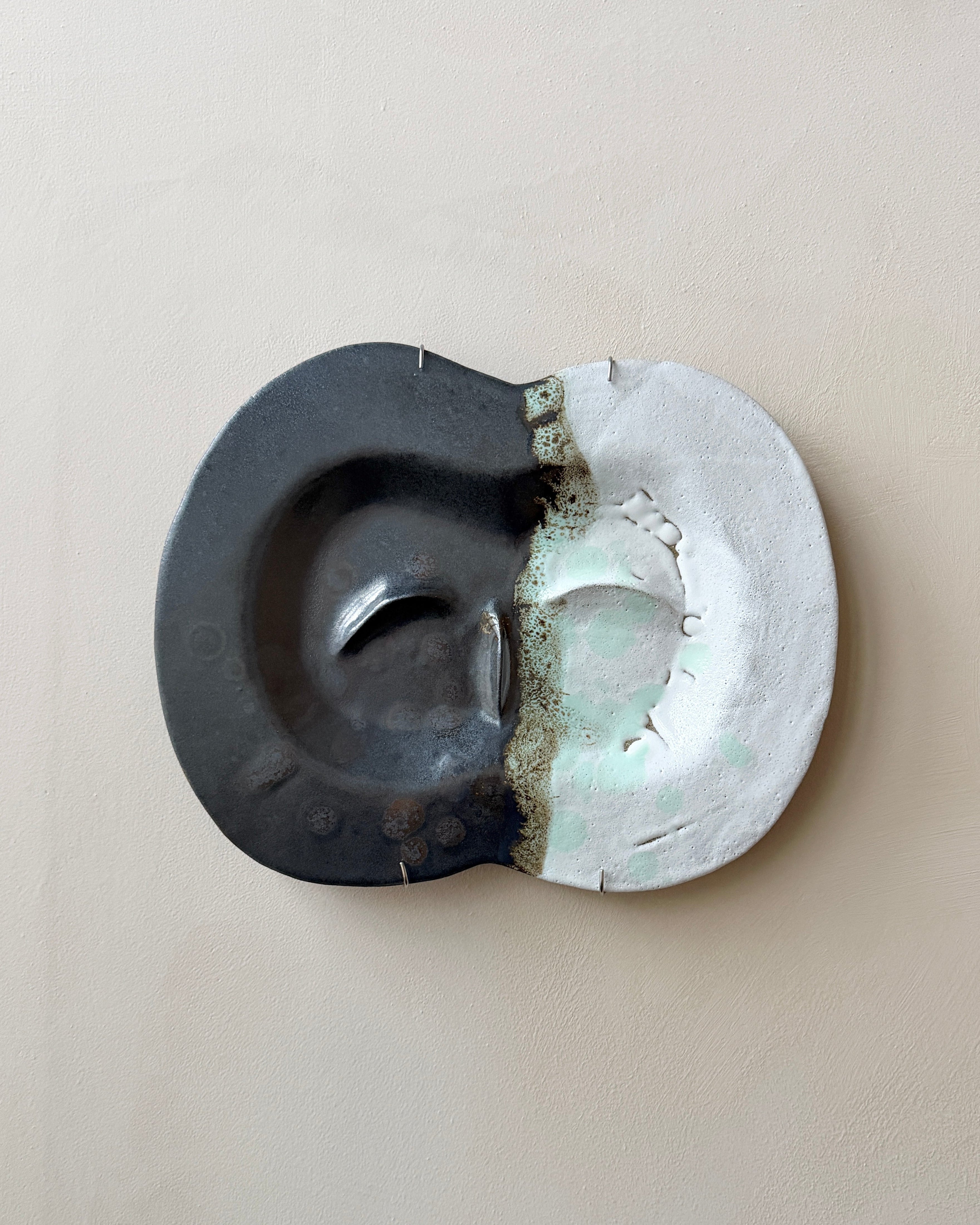 An abstract ceramic sculpture or decorative object with a split design, featuring a dark charcoal black left half and a matte white right half, separated by a textured, metallic bronze-gray seam. The object is photographed from directly above on a neutral, warm beige background.